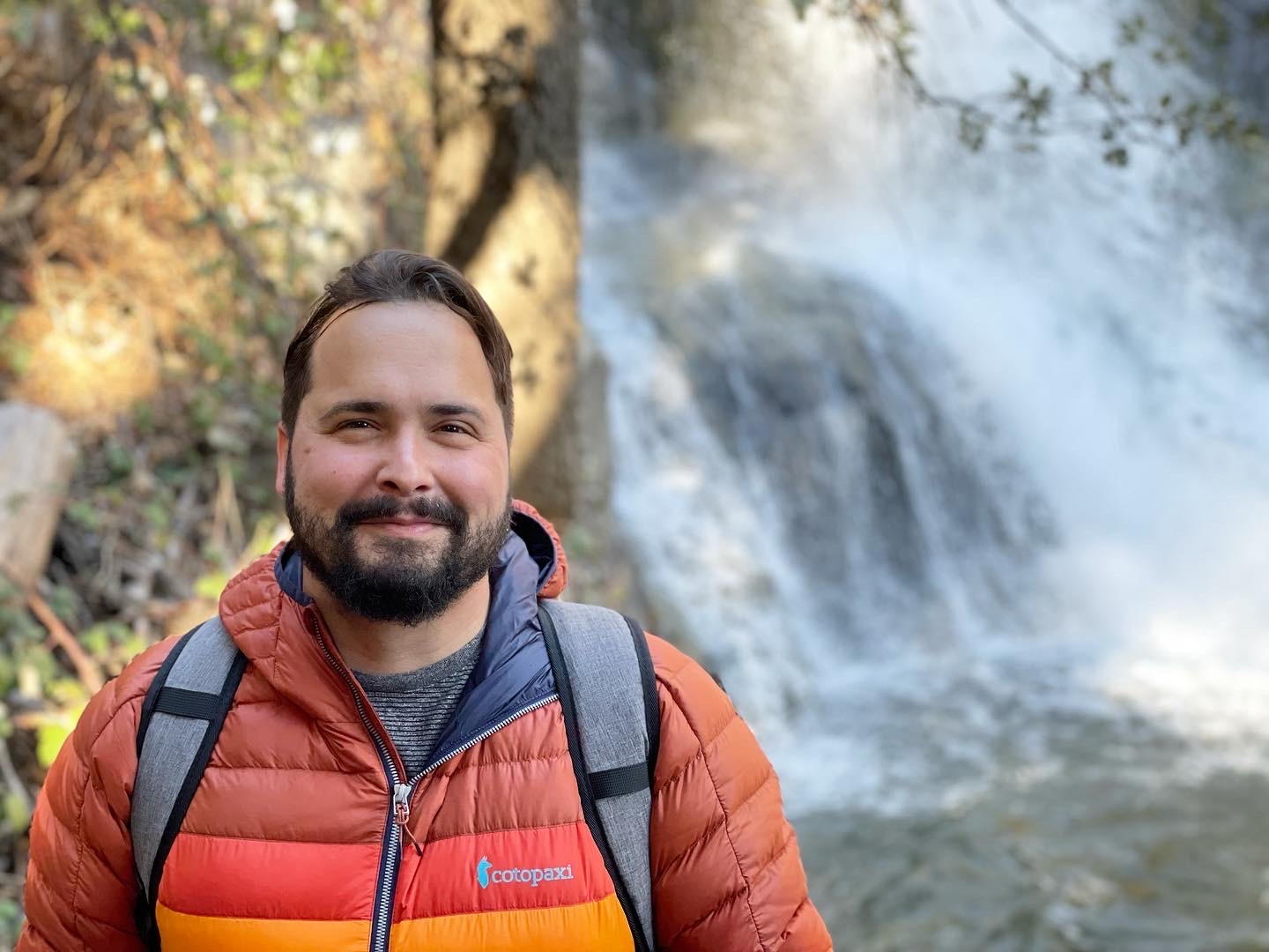 A photograph of a man wearing a red and orange jacket, smiling, and standing in front of a waterfall. The waterfall is cascading down in the background, surrounded by greenery and rocks. The man has a beard and short hair, and he is carrying a backpack. The overall scene is outdoors and captures the natural beauty of the waterfall and the serene environment.