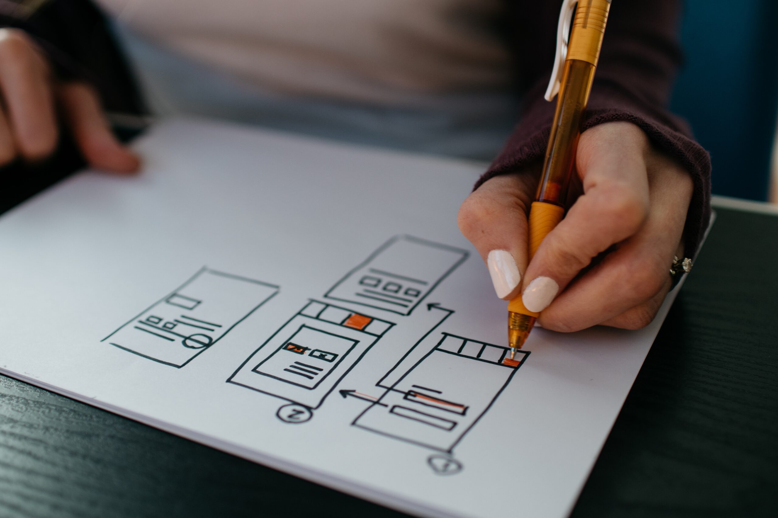 A close-up photograph of a person drawing a website wireframe on a piece of paper. The person’s hand is holding a pencil, sketching out the layout of a website with various sections and elements clearly marked. The image focuses on the hand and the paper, capturing the detailed process of planning and designing a website. The background is slightly blurred, indicating a focused and creative work environment.